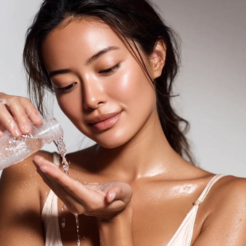 Femme très belle versant un toner liquide transparent dans sa main depuis un flacon neutre, dans une lumière douce et une ambiance spa haut de gamme. collection